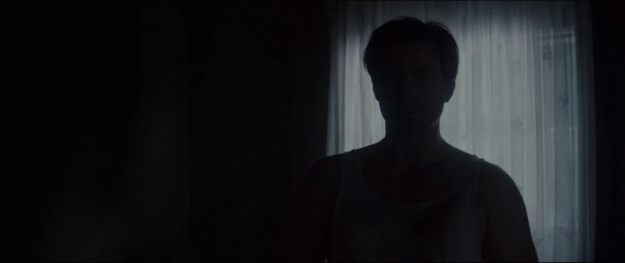 One & Two (2015) movie still: medium — A dimly lit interior room features a single window covered by a sheer white curtain. A man in a tank top…