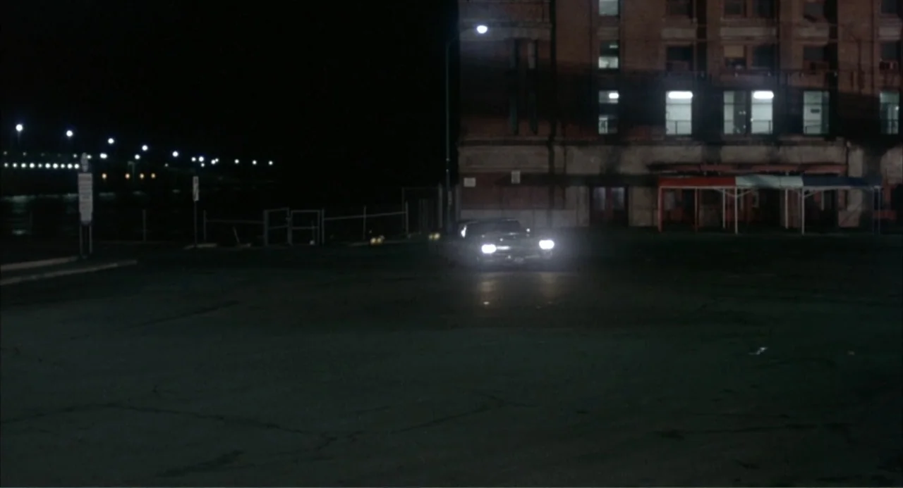 Cooley High (1975) movie still: wide — An empty asphalt parking lot sits before a dark brick industrial building at night. A lone vehicle with its…