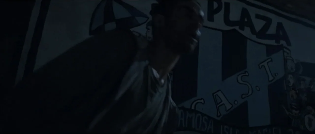 Gunman (2025) movie still: medium — A dark urban outdoor space features a concrete wall painted with a large emblem. A man passes by the wall in low…