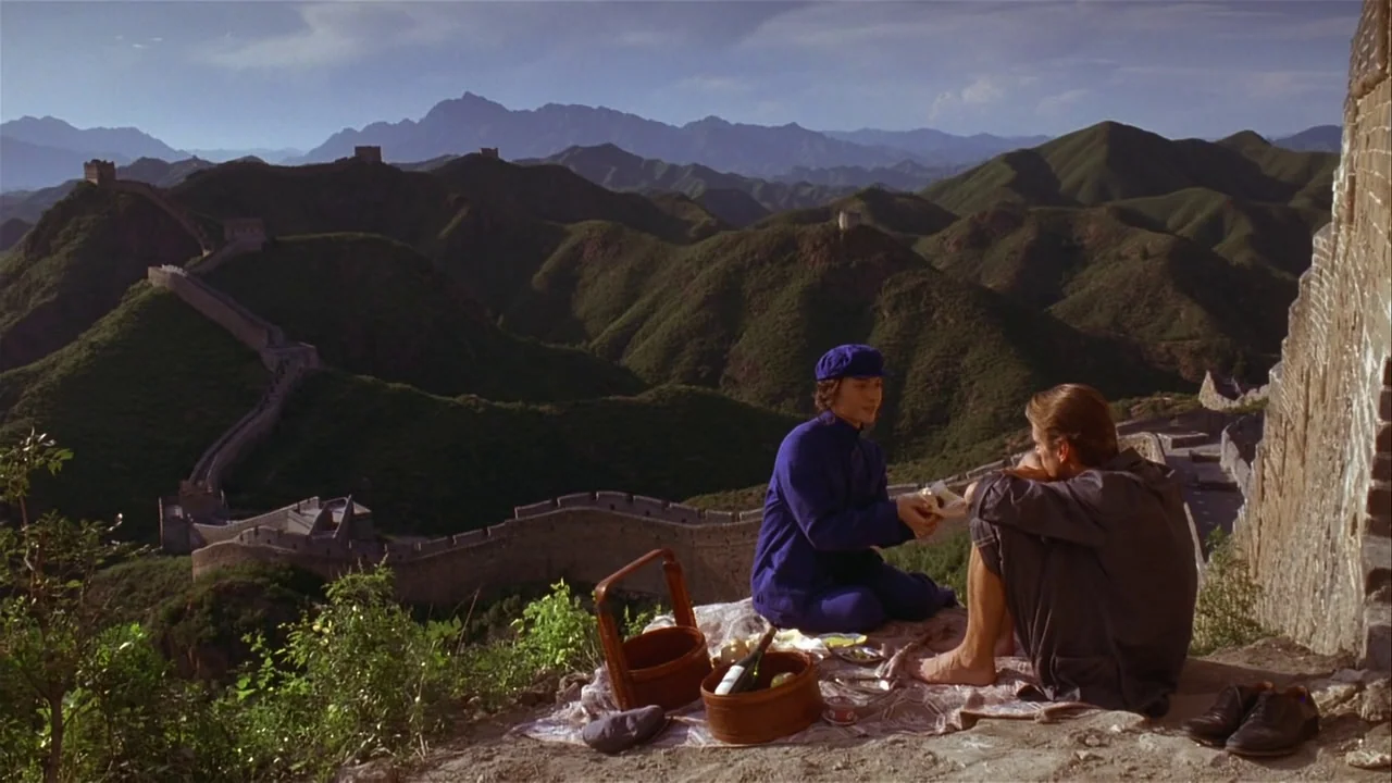 M. Butterfly (1993) movie still: wide — A man and a woman share a picnic atop a mountain path at the Great Wall. The historic structure stretches…