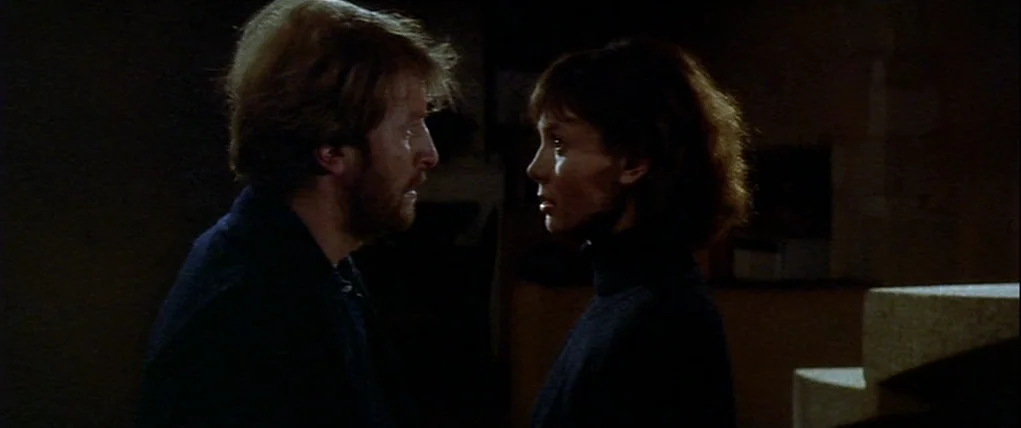 Love Unto Death (1984) movie still: medium — A dimly lit, shadowed room with two individuals standing close to each other. A bearded man and a woman…