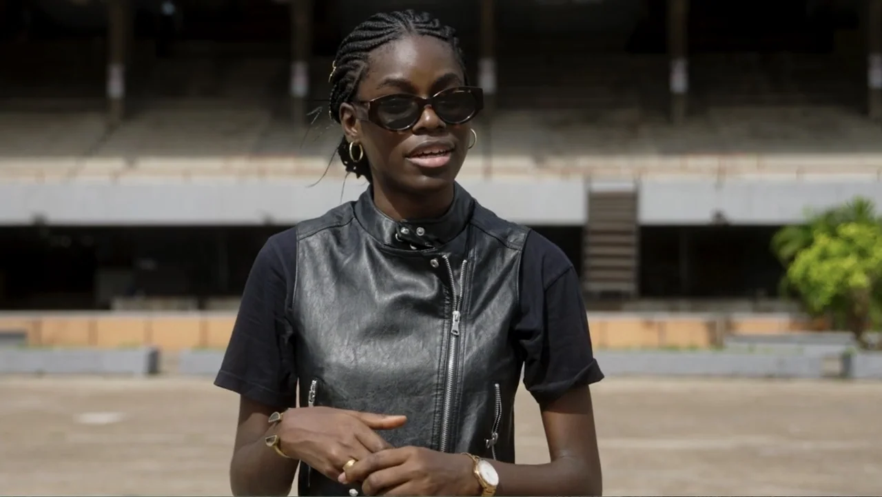 Victoria's Secret: The Tour '23 (2023) movie still: medium — A woman stands in the center of an empty concrete stadium concourse at daylight. The…
