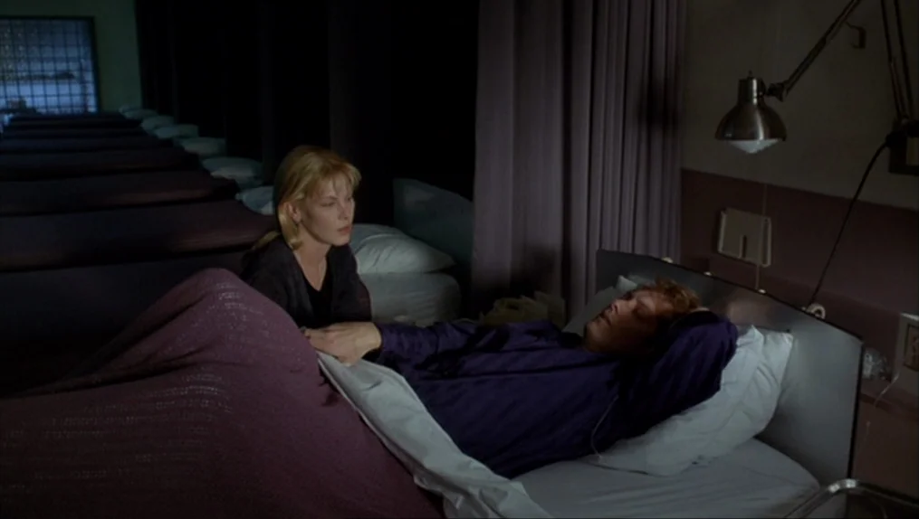 Crash (1996) movie still: medium — A dimly lit hospital room features a row of beds extending into darkness. A woman holds the hand of a man lying…