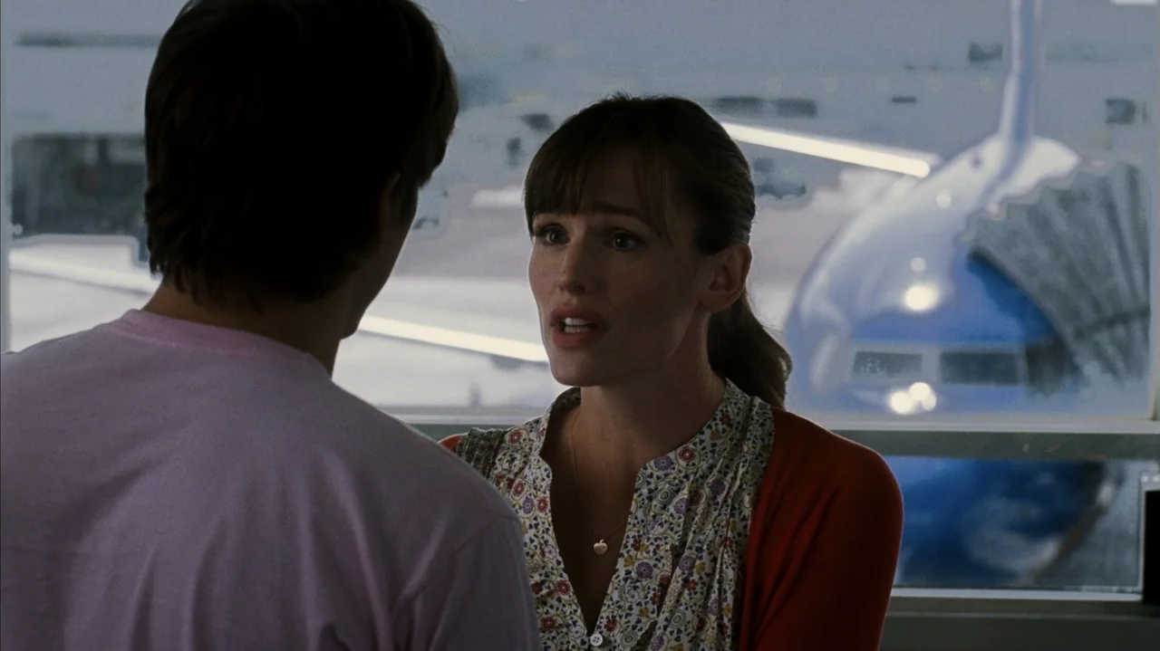 F Valentine's Day (2026) movie still: medium — An airport terminal interior features a large glass wall overlooking an airplane tarmac. A woman…