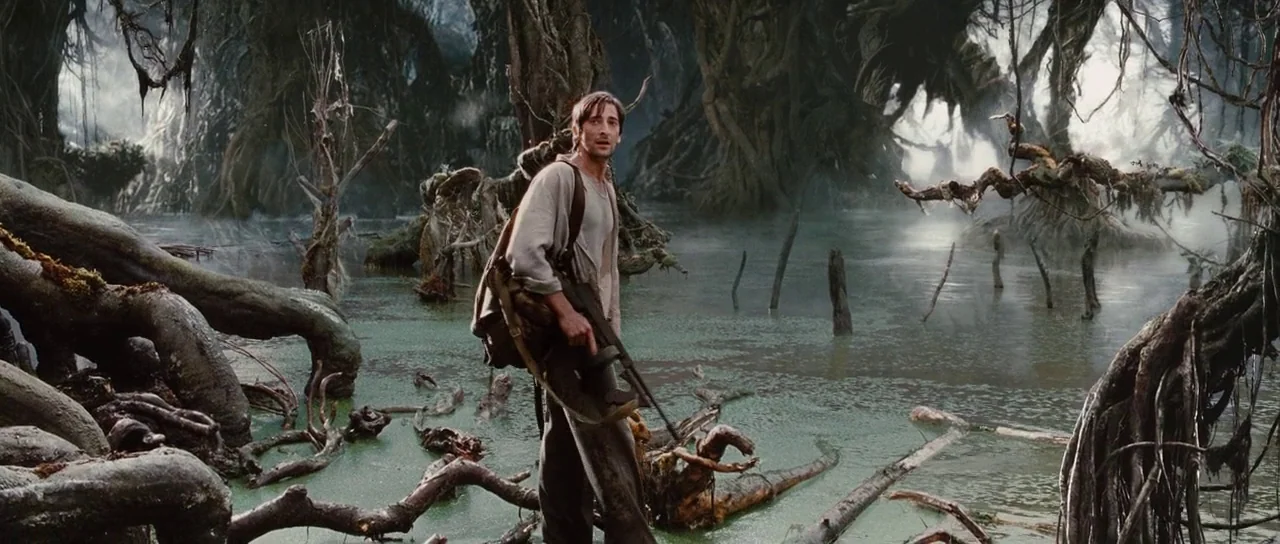 King Kong (2005) movie still: fullbody — A murky, water-logged landscape filled with thick, gnarled tree roots and dense vegetation. A man carrying…