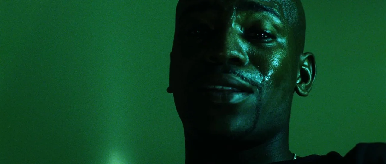 Dawn of the Dead (2004) movie still: closeup — A close-up shot focuses on a man against a dark, solid green background. Sweat beads glisten on his…