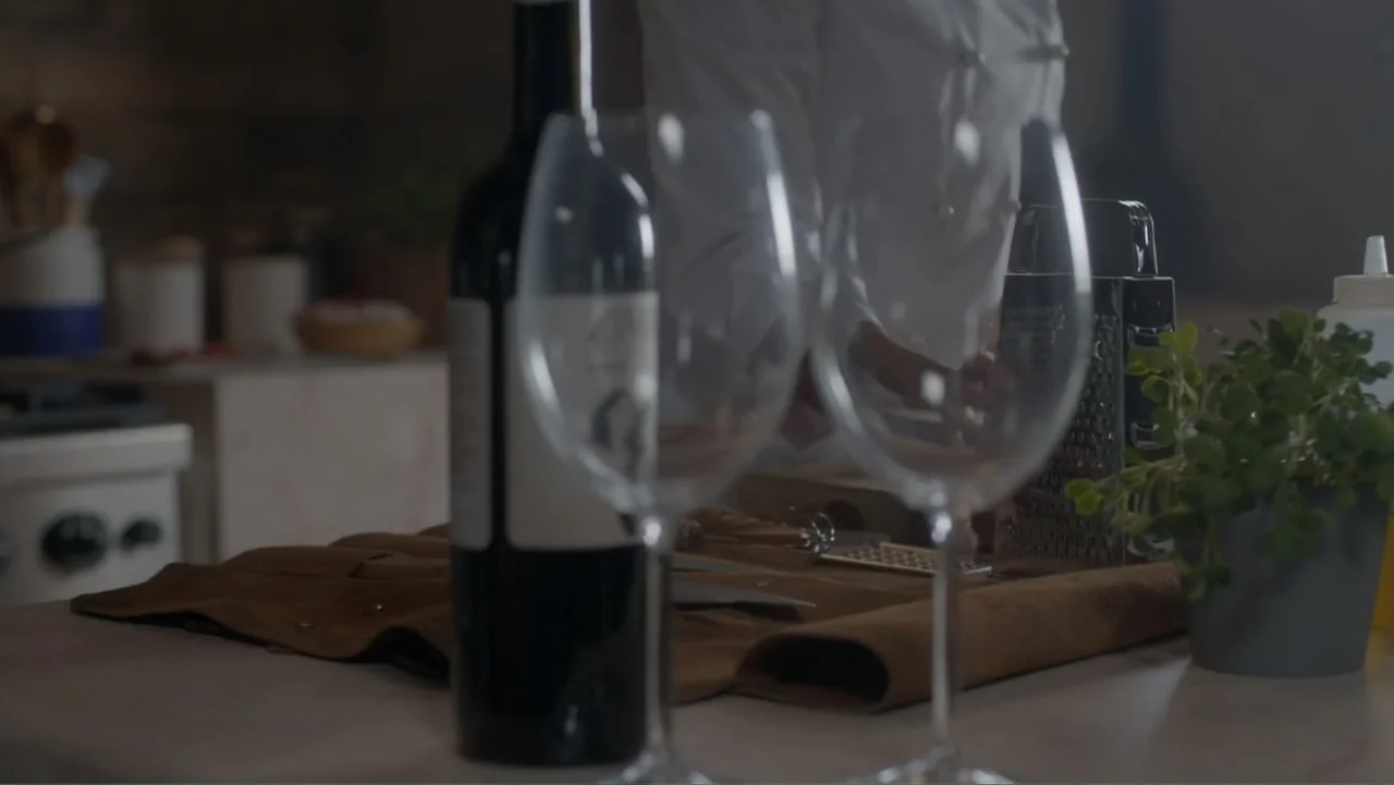 A Pinch of Portugal (2023) movie still: medium — A professional kitchen counter holds a red wine bottle and two clear wine glasses. A chef wearing a…