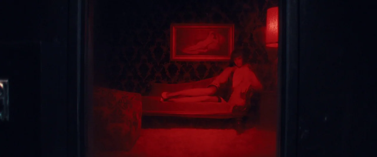MaXXXine (2024) movie still: wide — A dark, red-saturated room contains a patterned chaise lounge and a framed painting. A woman with dark hair…