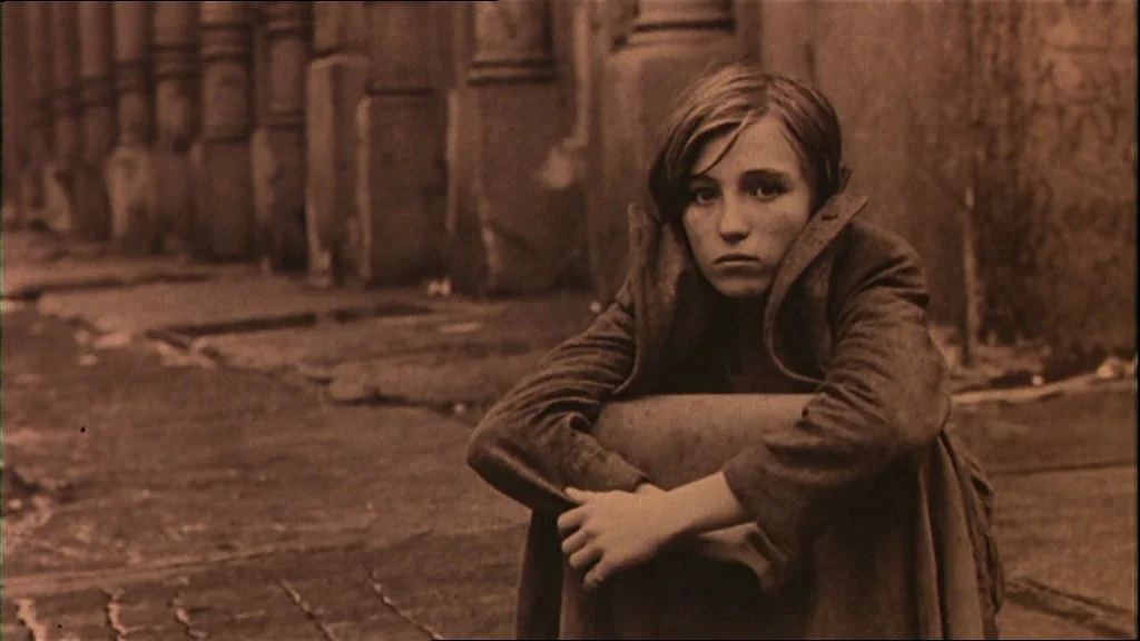 Days Of Heaven (1978) movie still: medium — A stone paved urban street with blurred architectural columns in the background. A young girl in an…