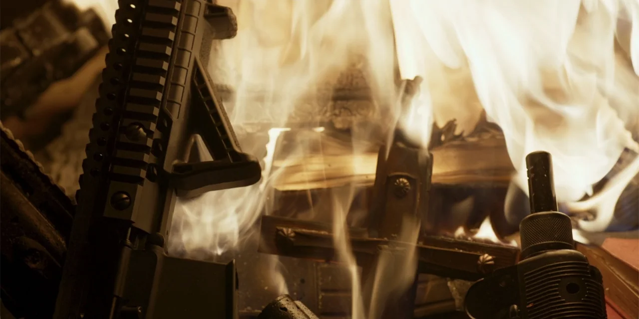 Demonic (2015) movie still: closeup — A close view of a fire burning inside a stone or brick hearth. A black rifle and a small metal cross rest…