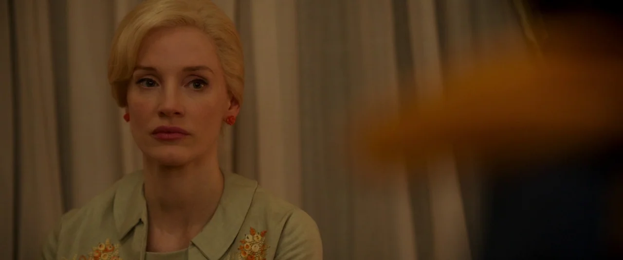 Mother’s Instinct (2024) movie still: closeup — A woman with styled blonde hair sits indoors in front of fabric curtains. She gazes off-frame at an…