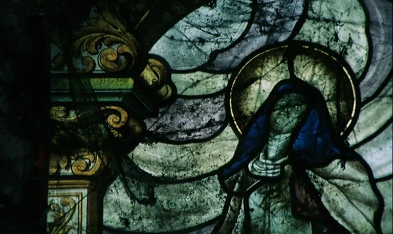 The Rose King (1986) movie still: closeup — A close-up view shows a weathered stained glass panel featuring an ornamental border. A stylized, haloed…