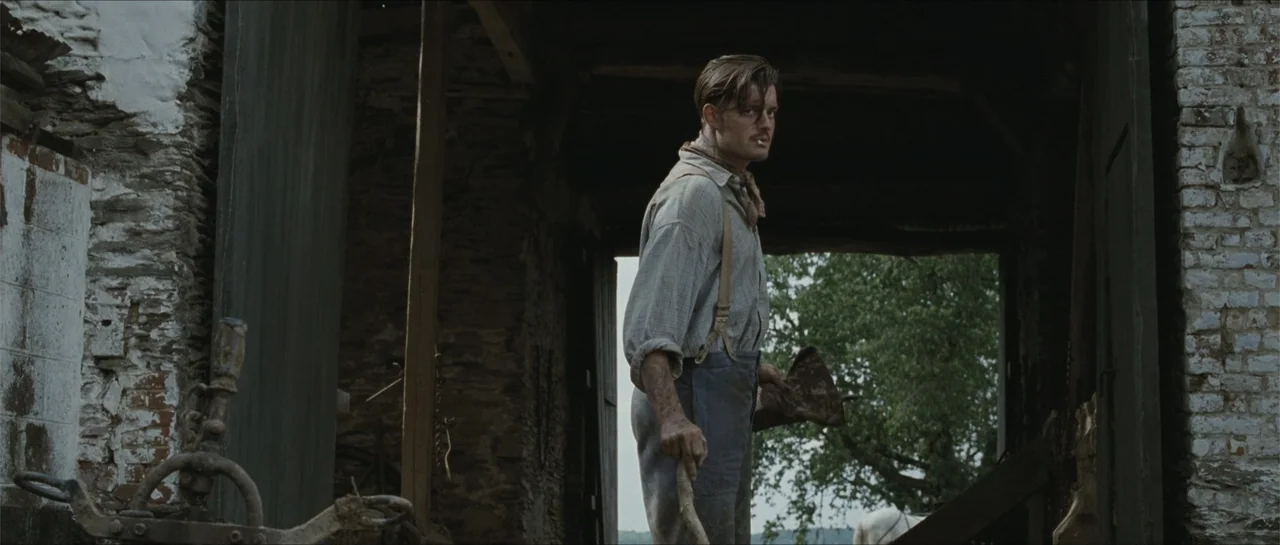 Suite Française (2015) movie still: medium — A rustic barn interior features stone walls and an open doorway leading outside. A young man in period…