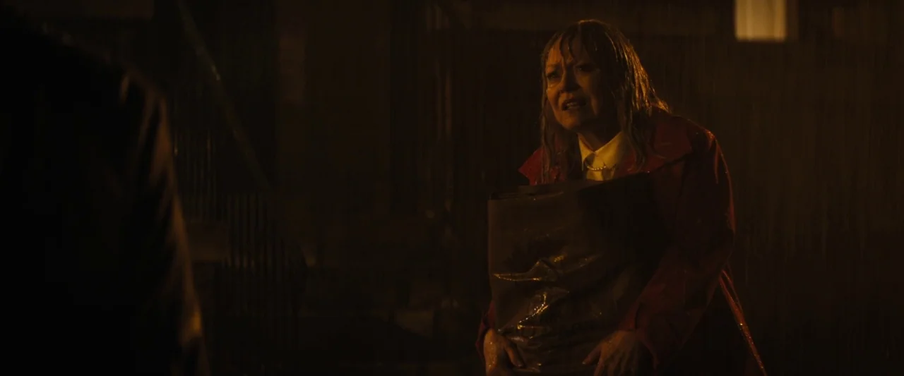 The Grudge (2020) movie still: medium — A dark outdoor urban setting at night is illuminated by heavy rain. A woman in a red coat stands in the…