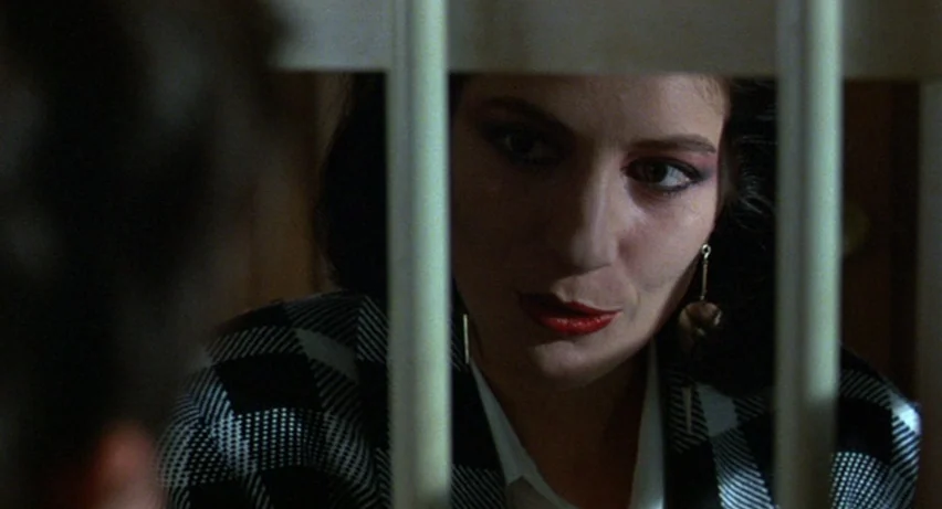 Matador (1986) movie still: closeup — A dimly lit room features white vertical iron bars dividing the frame. A woman with dark hair looks towards an…
