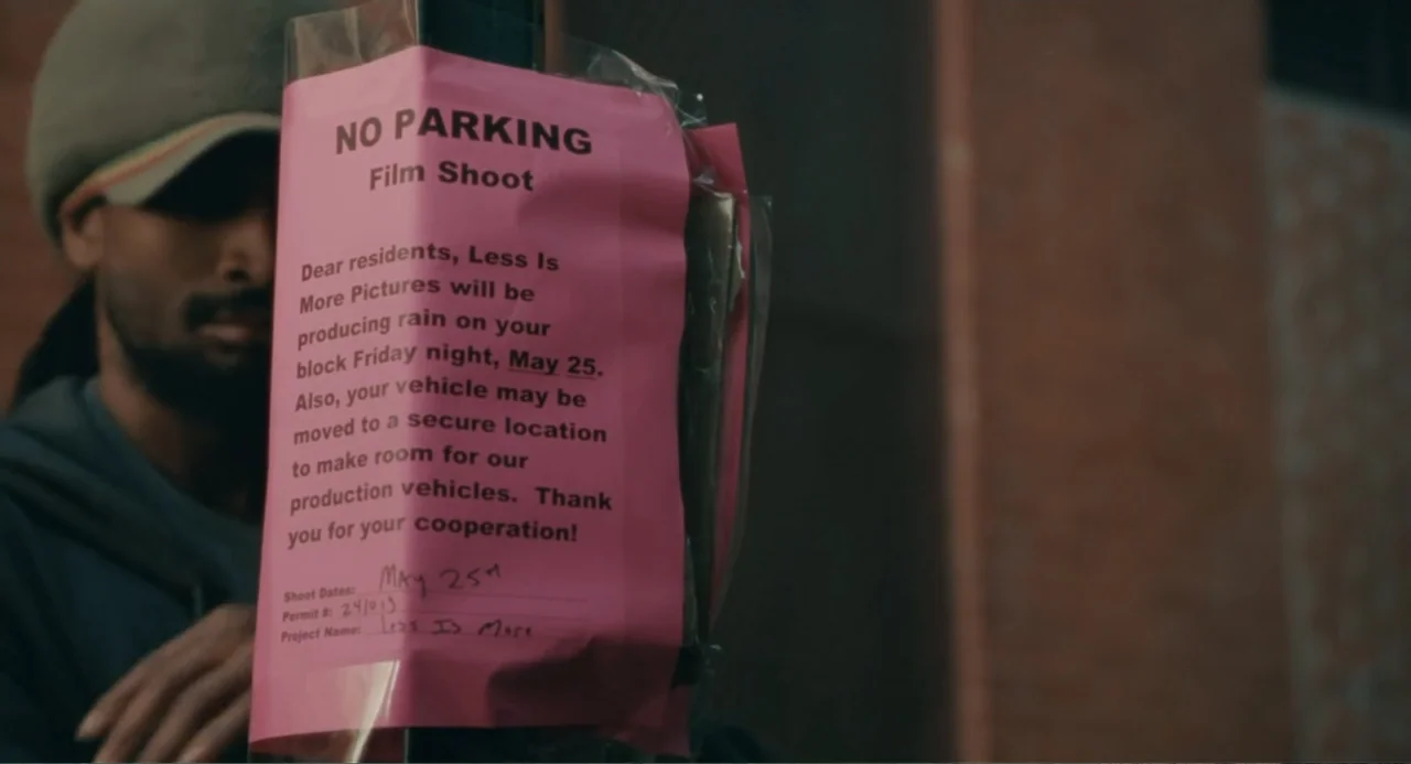Motherhood (2020) movie still: closeup — A pink film shoot parking notice is attached to a metal post outdoors. A man in a hooded sweatshirt walks…