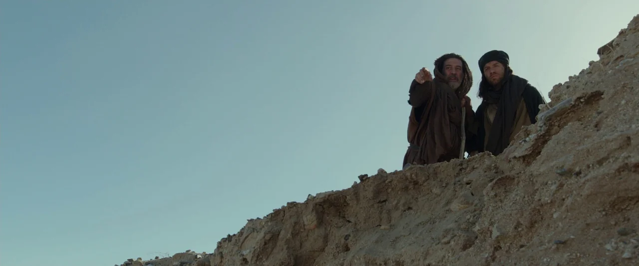 Last Days in the Desert (2015) movie still: wide — An arid, elevated dirt bank stands out against a clear, pale blue sky. Two men in ancient-style…