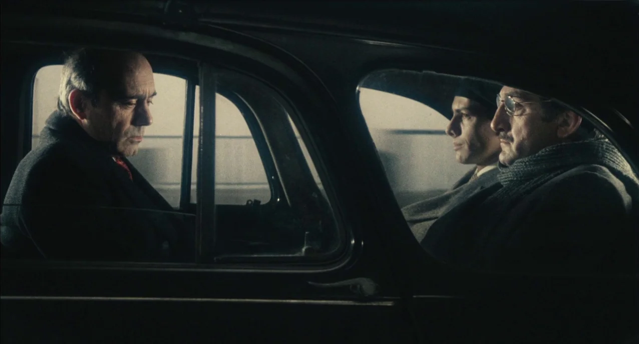 The Army of Shadows (1969) movie still: medium — Three men sit inside the back of a vintage car during the day. The men remain still as the vehicle…