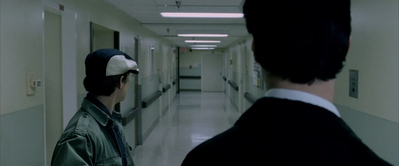 Constantine (2005) movie still: medium — A long, sterile hospital hallway features beige walls and industrial ceiling lights. Two men stand in the…