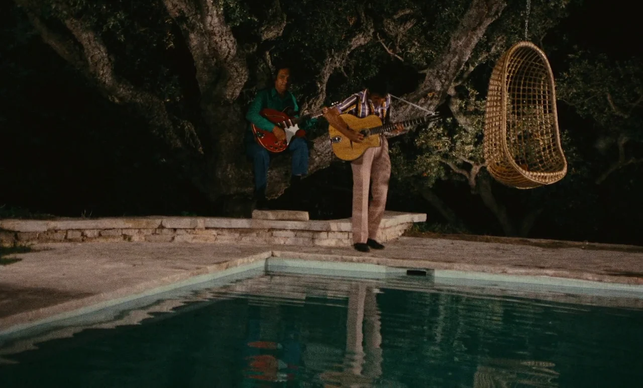 The Swimming Pool (1969) movie still: wide — An outdoor paved area borders a swimming pool at night beneath a large oak tree. Two men play guitars…