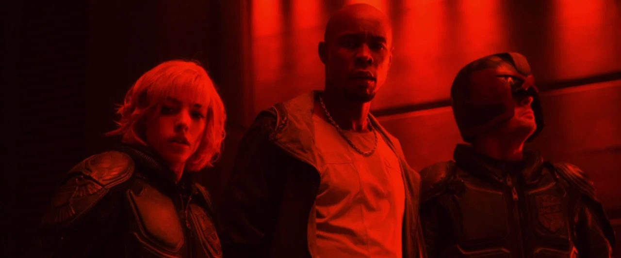 Dredd (2012) movie still: medium — Three people stand in a dim, futuristic hallway bathed in intense red light. They look toward the camera with…