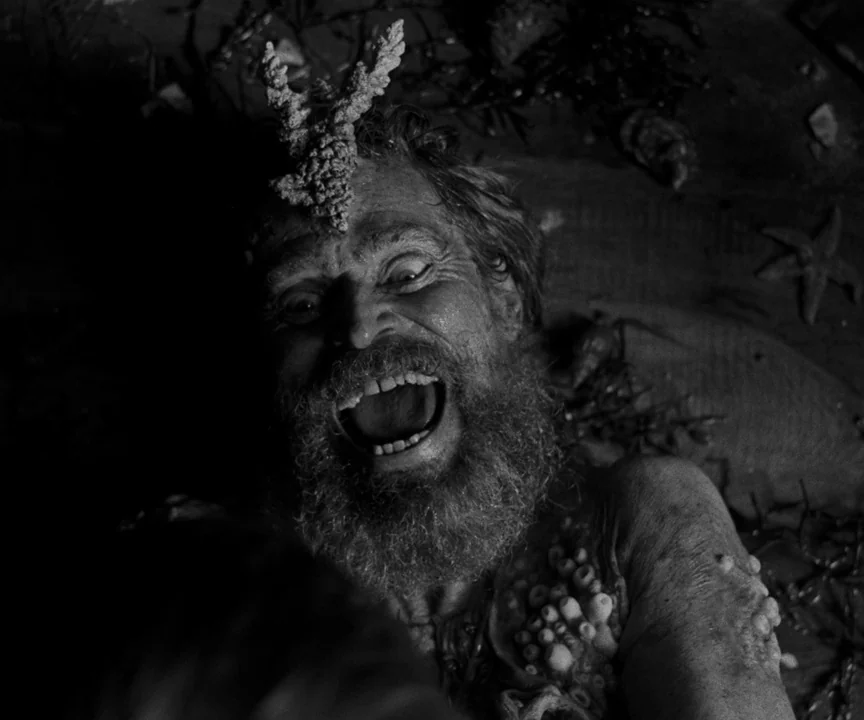 The Lighthouse (2019) movie still: closeup — An elderly man lies on wet sand covered in barnacles and sea growth. He looks upward with a…