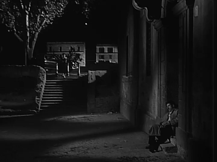 Variety Lights (1950) movie still: wide — A dark urban environment at night featuring stone buildings and an empty staircase. A man sits in a…