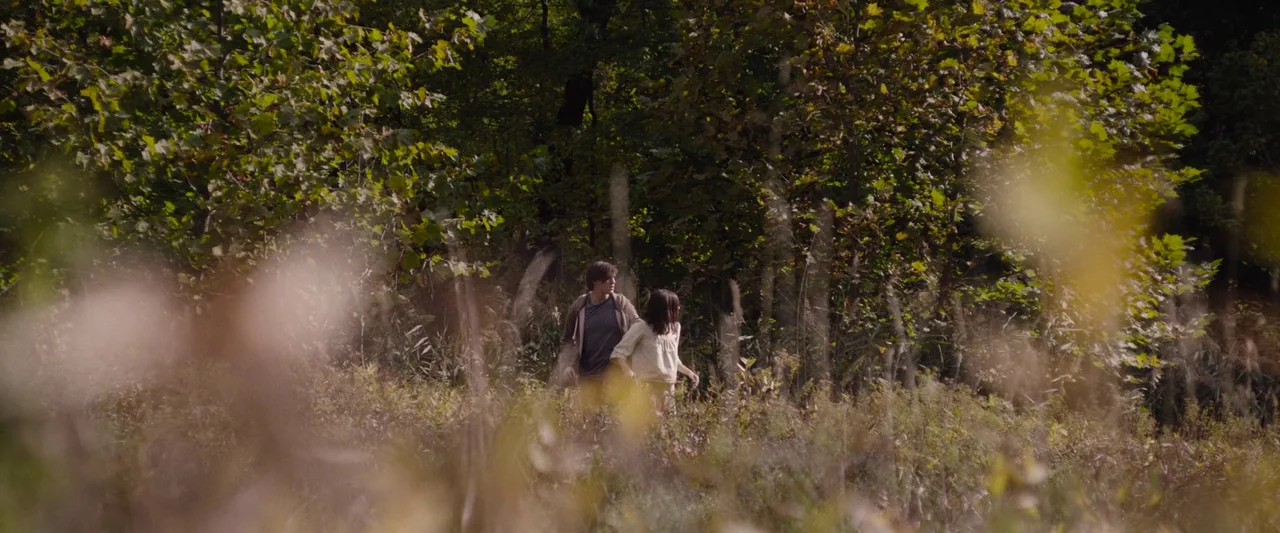Wildling (2018) movie still: wide — An open grassy area leads into a dense line of mature deciduous trees. A young man and woman walk together…