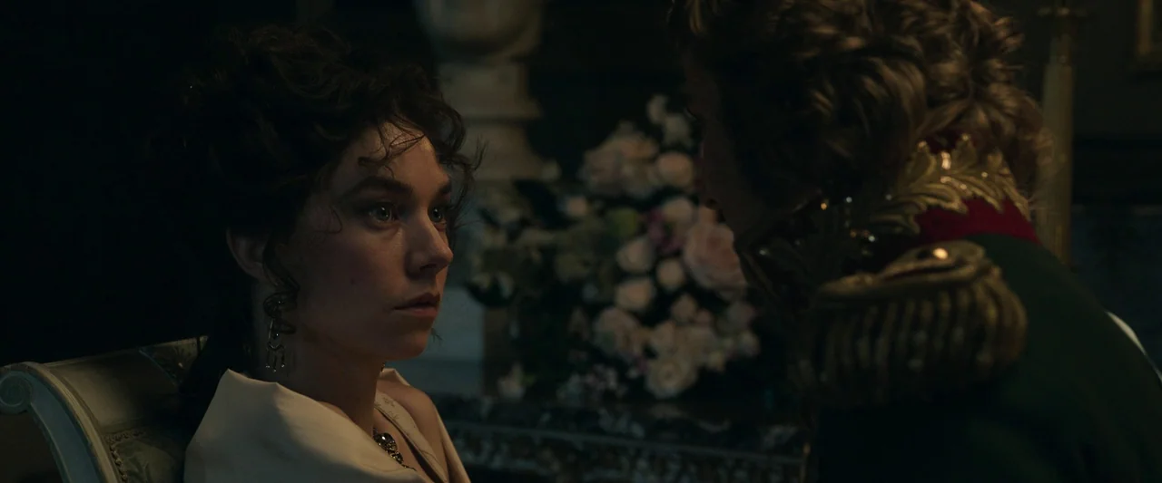 Napoleon (2023) movie still: medium — An opulent 19th-century interior space features ornate woodwork and a bouquet of flowers. A woman faces an…