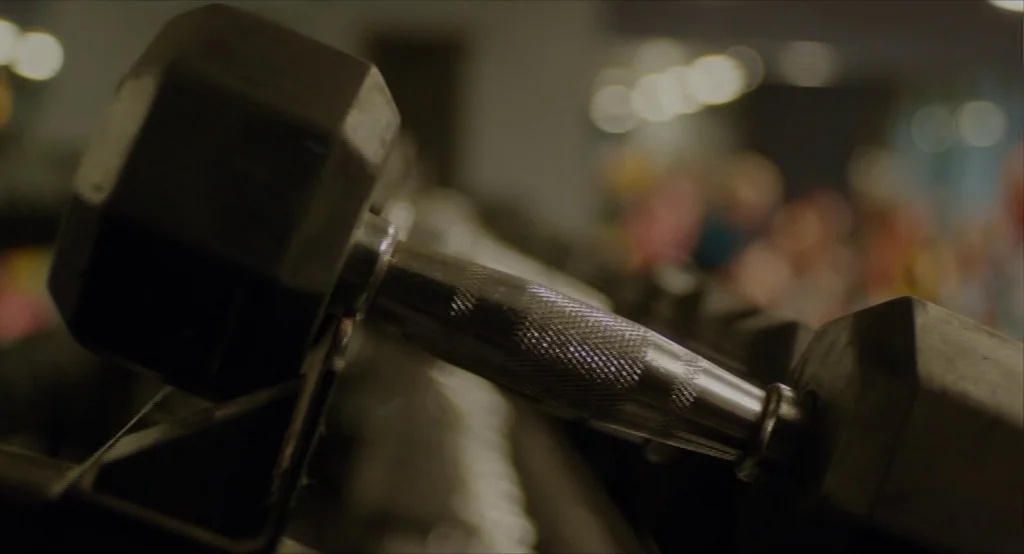 Results (2015) movie still: closeup — A close-up view shows a black rubber-coated hexagonal dumbbell resting on a metal rack. The knurled steel…