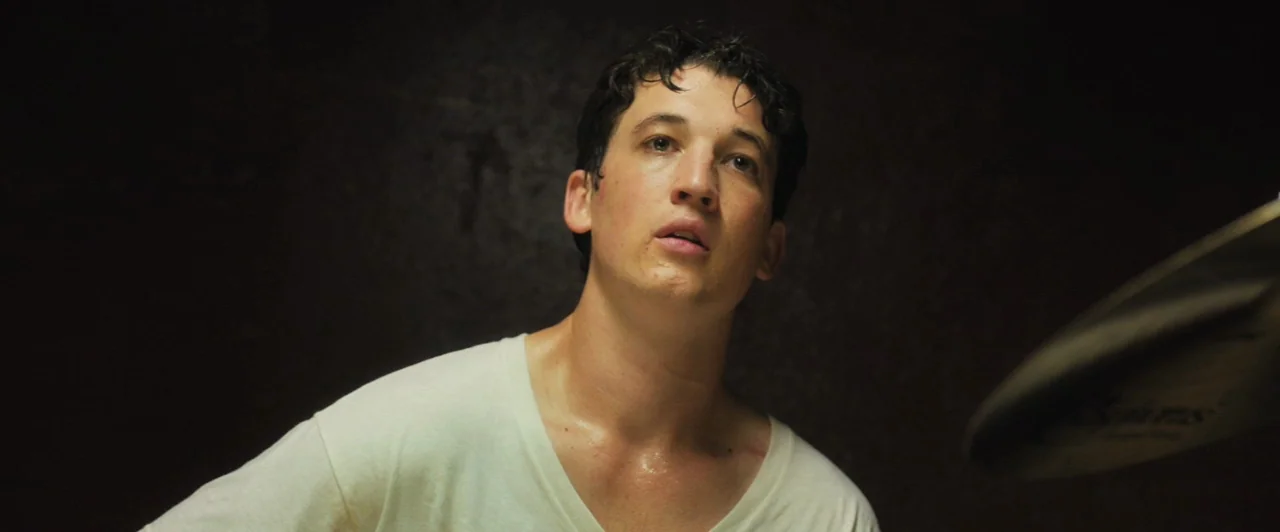 Whiplash (2014) movie still: closeup — A dark practice space with a solid brown background and minimal visible objects. A young man wearing a white…