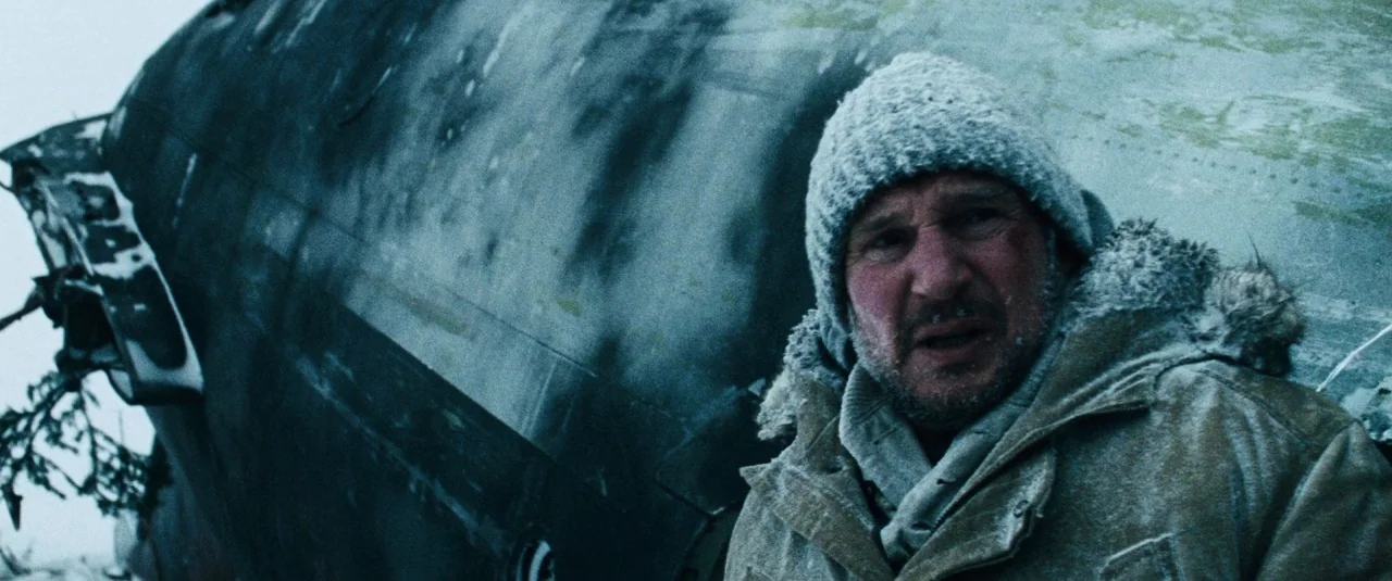 The Grey (2012) movie still: medium — A man stands outdoors near the large metal hull of a crashed airplane. The surface of the plane is dark and…