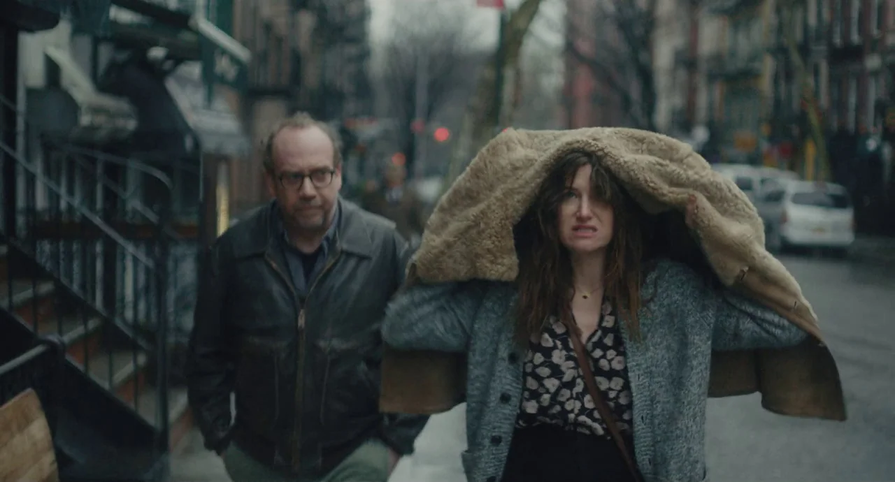 Private Life (2018) movie still: medium — An urban sidewalk lined with brick townhouses on a cloudy day. A man and a woman walk towards the camera…
