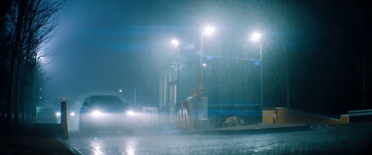 Venom (2018) movie still: wide — A glass security booth stands illuminated by bright floodlights amidst a heavy downpour. A dark car moves slowly…