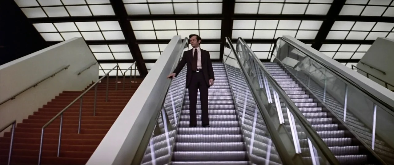 The Parallax View (1974) movie still: fullbody — A man stands on an escalator beneath a bright grid of overhead lights. The escalator is flanked by…