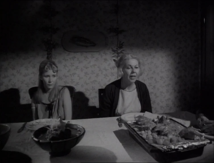 Eraserhead (1977) movie still: medium — A dimly lit room with patterned wallpaper features a table set for dinner. Two women sit facing forward…