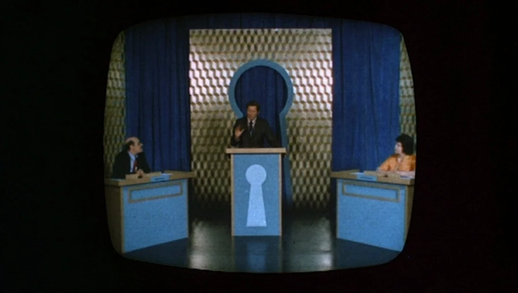 Sisters (1973) movie still: wide — A 1970s television game show set features blue podiums and a keyhole cutout background. A man stands at the…