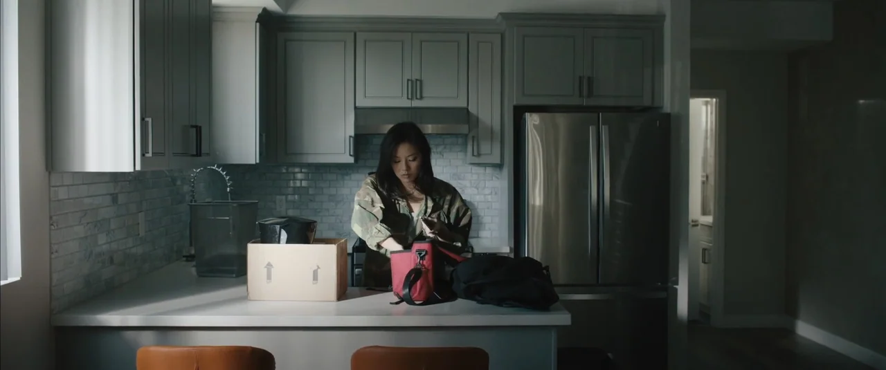 Stalker (2020) movie still: medium — A modern kitchen with grey cabinetry and white countertops contains a cardboard box. A woman in a camouflage…