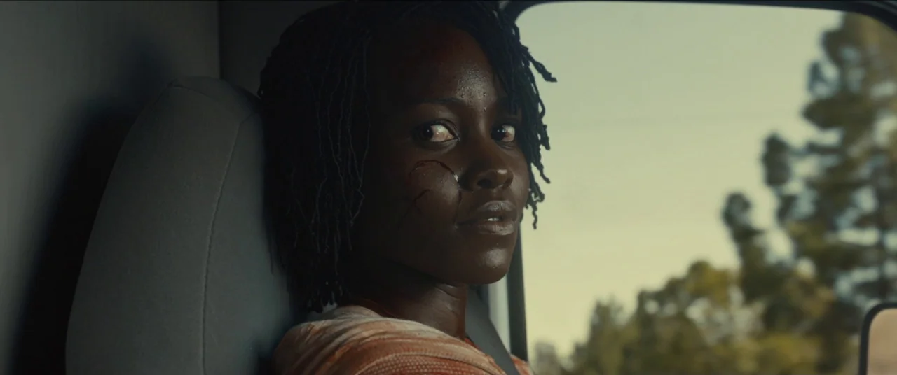 Us (2019) movie still: closeup — A dark, cramped interior of a vehicle passenger seat with a gray headrest visible. A woman with a facial injury…