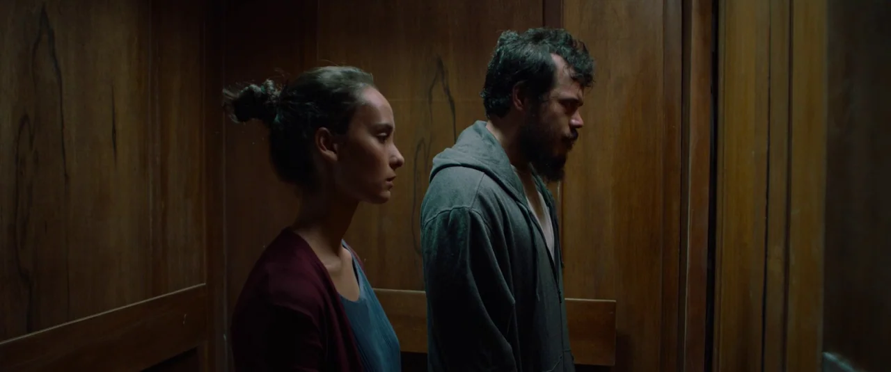 The Night Comes for Us (2018) movie still: medium — The interior of an elevator is paneled in uniform vertical wooden slats. A man and woman stand…
