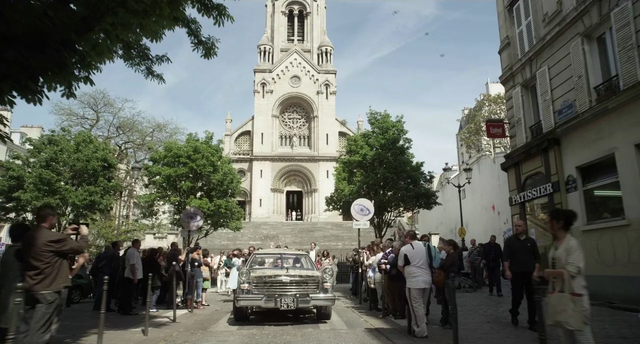 Mood Indigo (2013) movie still: wide — A crowd lines a cobblestone street in front of a stone church with steps. A vintage car drives toward the…
