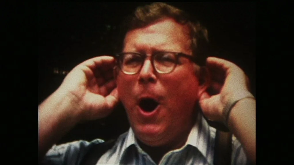 Junebug (2005) movie still: closeup — An outdoor setting with a dark, blurred background and indistinct vegetation. A man in eyeglasses holds both…