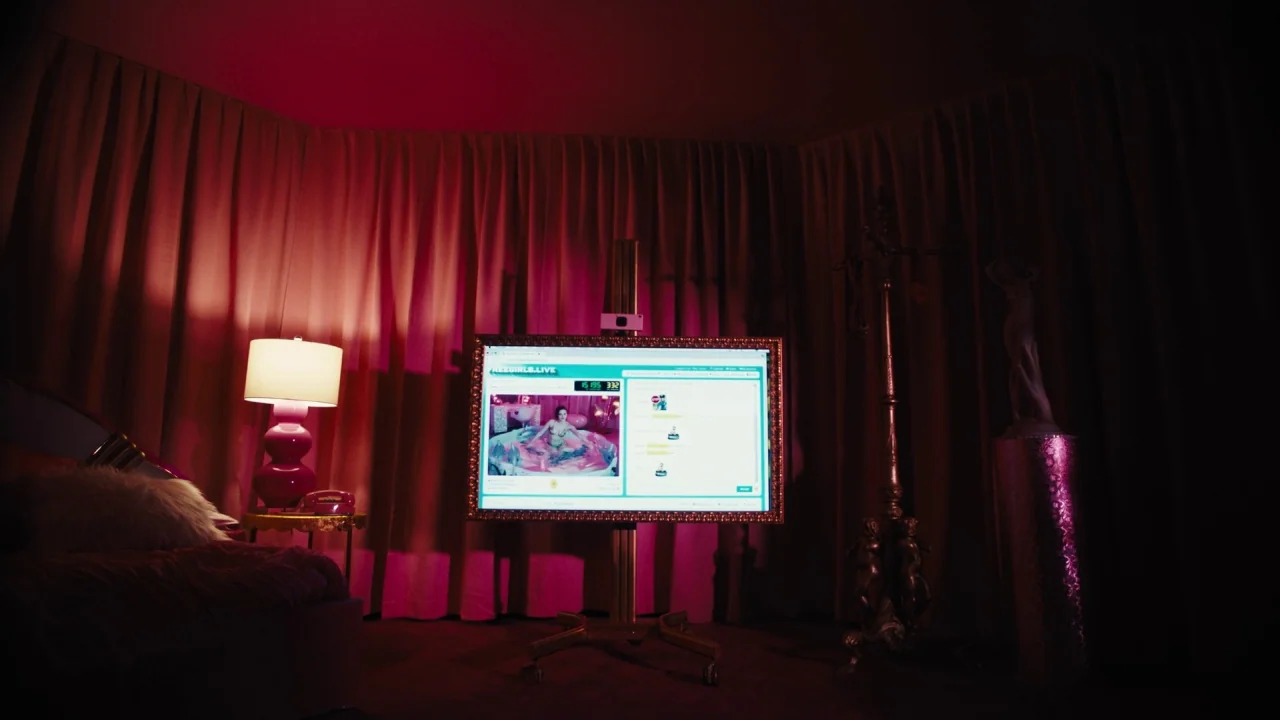 Cam (2018) movie still: wide — A dimly lit room features heavy pink curtains and a large monitor on wheels. A webcam displays a live stream on the…