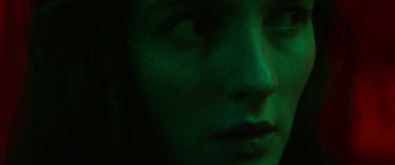 Kissing Candice (2017) movie still: closeup — A dark, unidentifiable space is split between harsh green and red light sources. A young woman looks…