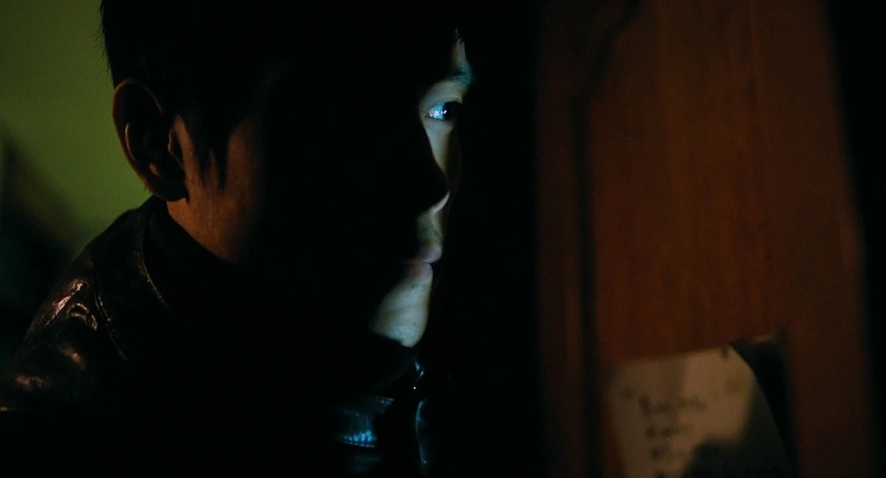 I Saw The Devil (2010) movie still: closeup — A dark, dimly lit indoor space with a wooden surface on the right. A man in a dark jacket is…