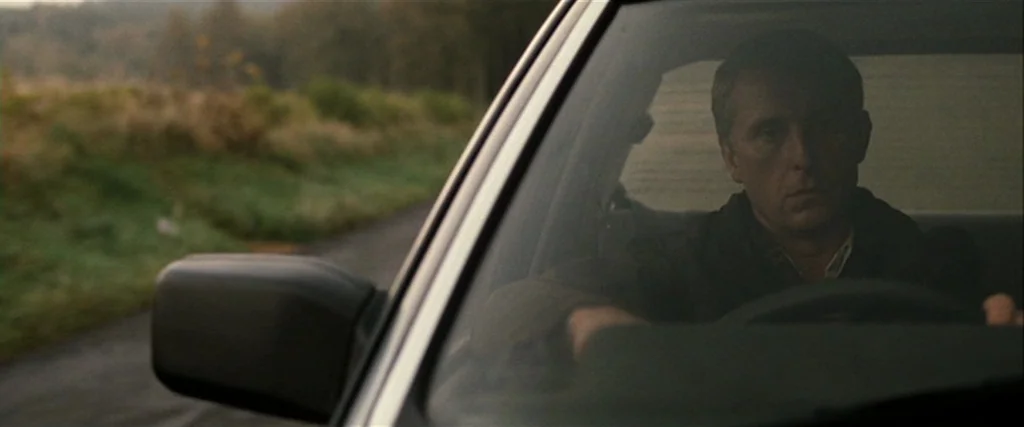 Hunger (2008) movie still: medium — A man sits in the driver seat of a car parked on a rural road. The windshield frames his upper body against a…