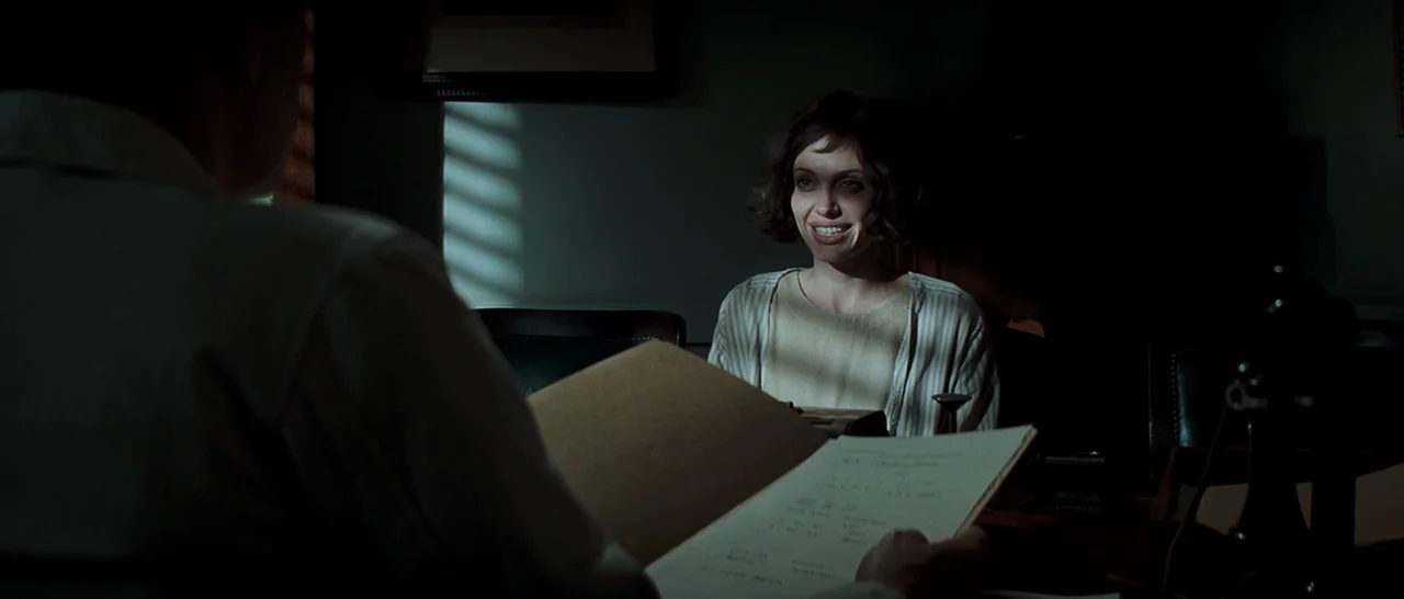 Changeling (2008) movie still: medium — A dark, sparsely furnished room features Venetian blinds casting light stripes on the walls. A woman sits at…