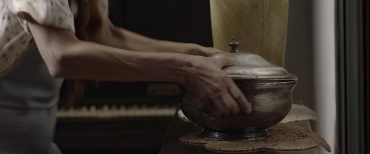 Like or Die (2023) movie still: closeup — A domestic interior features a wooden surface supporting a decorative metallic lidded bowl. A person…