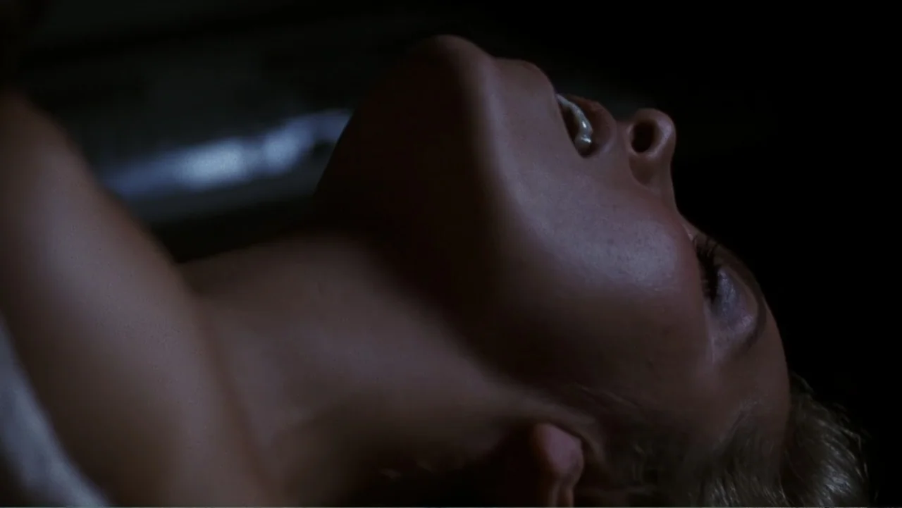 Presumed Innocent (1990) movie still: closeup — A dark, nondescript interior space features a close view of a person lying back. A woman with blonde…