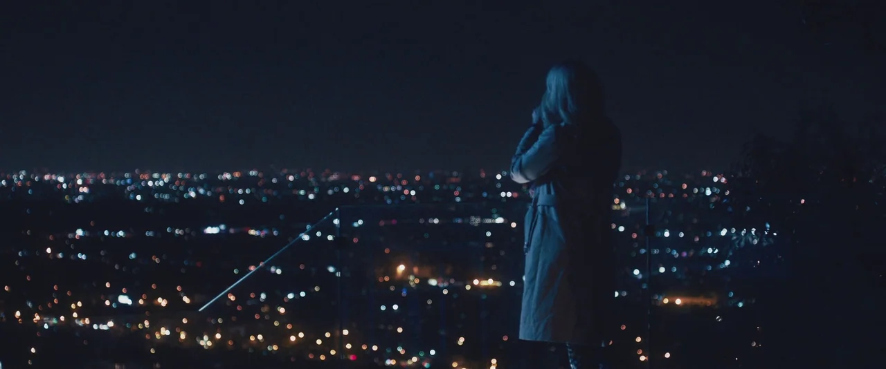 Gemini (2017) movie still: fullbody — A dark outdoor lookout area overlooks an expansive, blurred city night skyline. A blonde woman in a light…